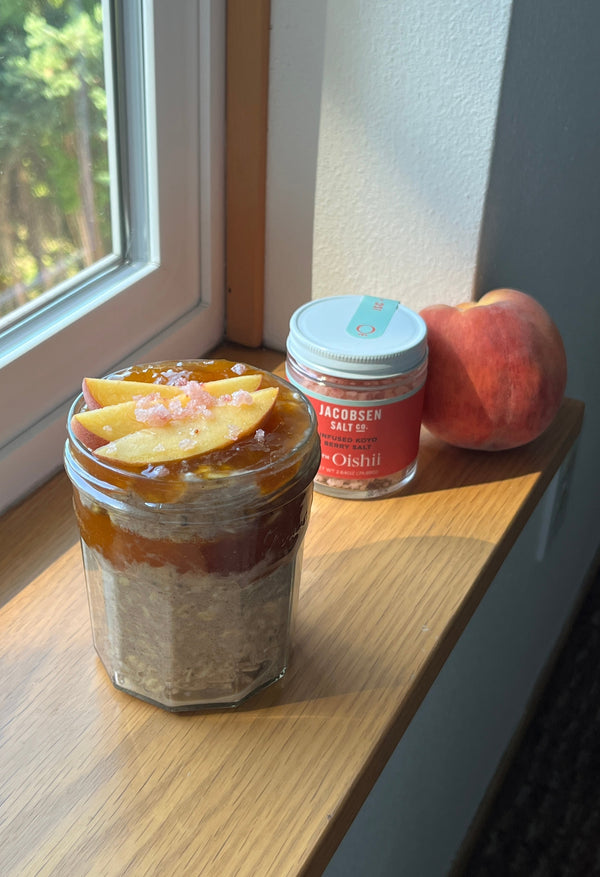 Cup of overnight oats in a glass, topped with sliced peaches and Jacobsen Koyo Berry Salt on a window sill.