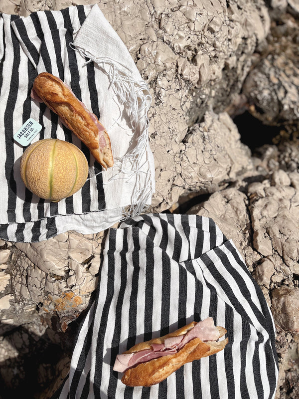 Two beach towels on rocks. On top of the beach towels are baguette sandwiches, a melon, and a sea salt slide tin.
