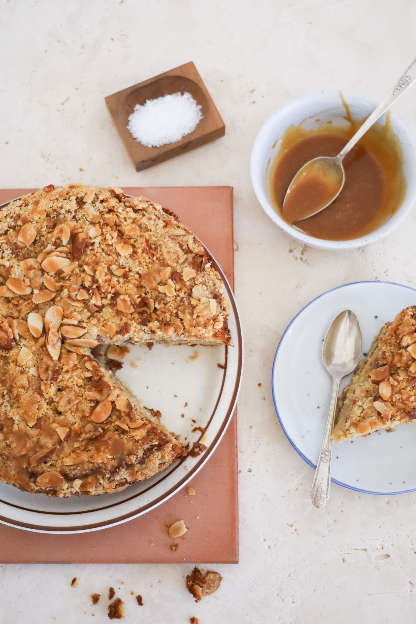 Cake covered with almond topping with one triangular cut out. The slice plated and off to the right, next to a bowl of caramel sauce and a salt cellar.