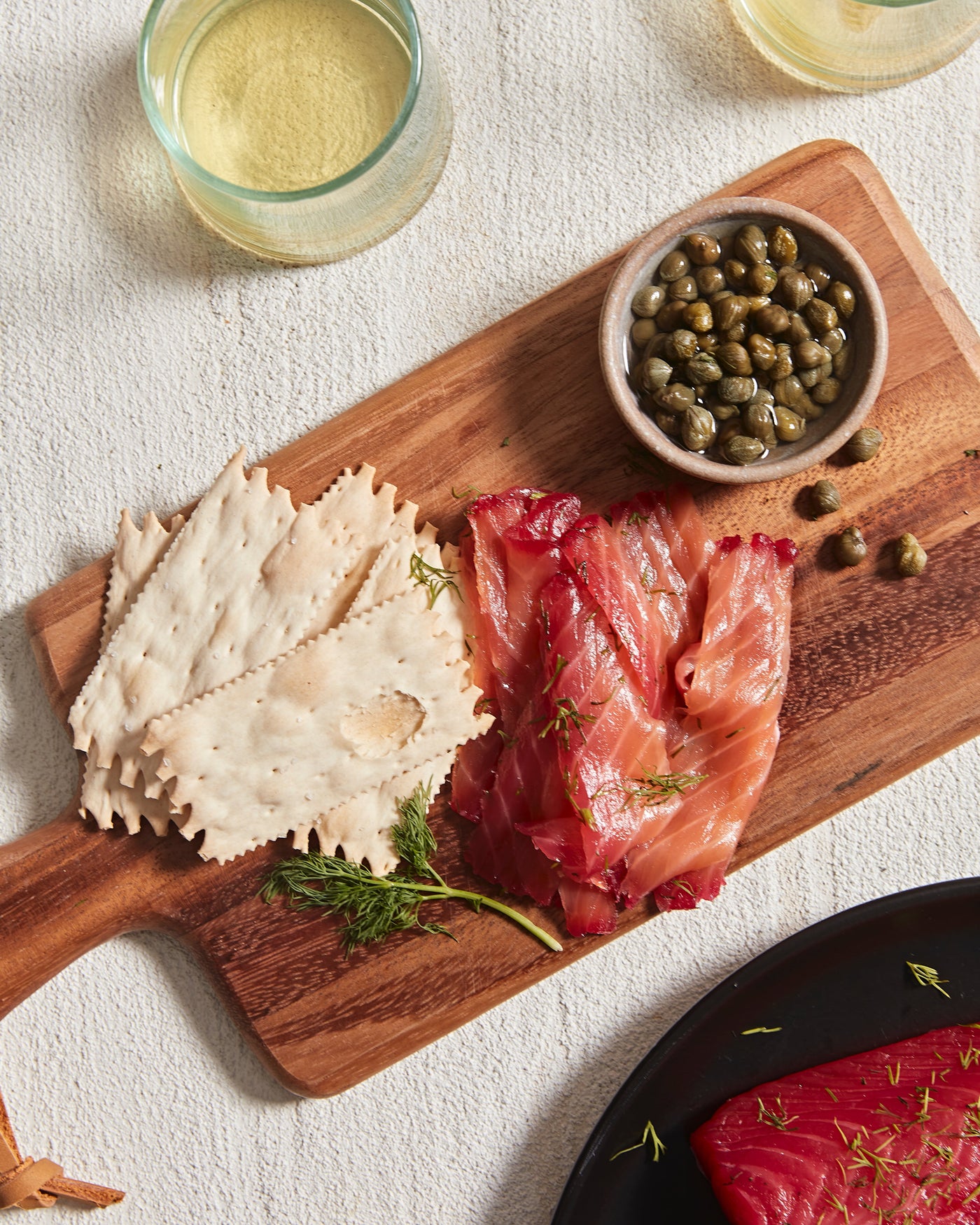 A wood cutting board with crackers, beet-cured salmon gravlax, and a bowl of capers.