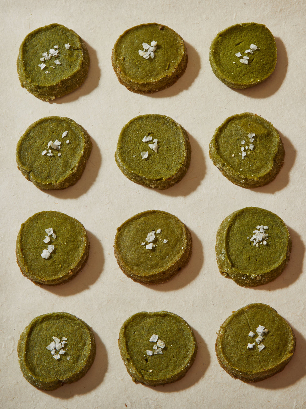 A dozen round green cookies, each topped with flake salt.