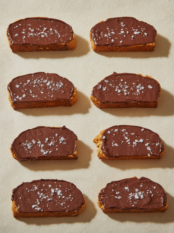 Eight dark brown biscotti cookies, topped with dark chocolate spread and a sprinkle of sea salt on top.