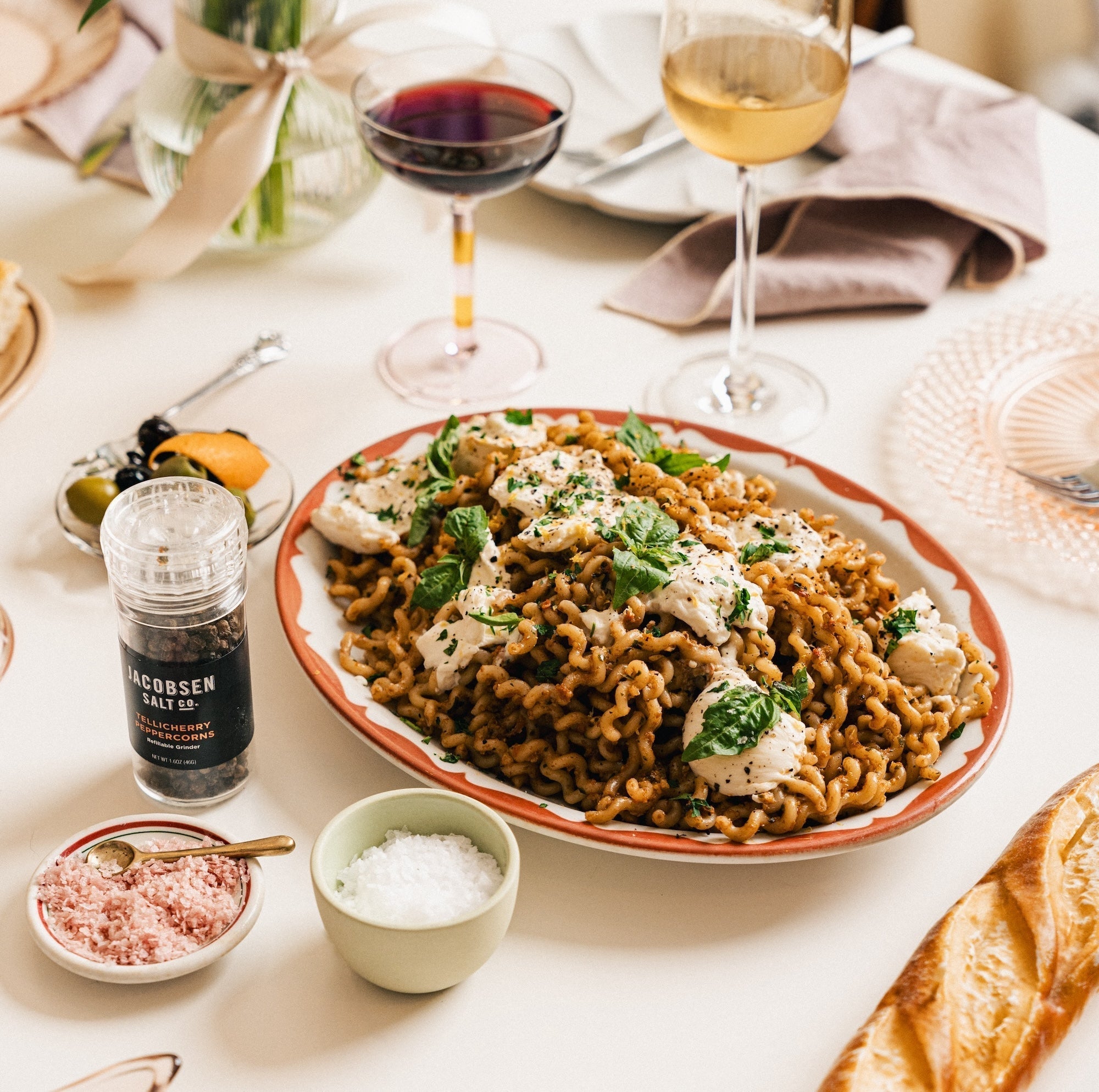 Dark brown pasta topped with burrata cheese and green basil plated on a fancy table top with glasses of wine, a black pepper grinder and salt cellars.