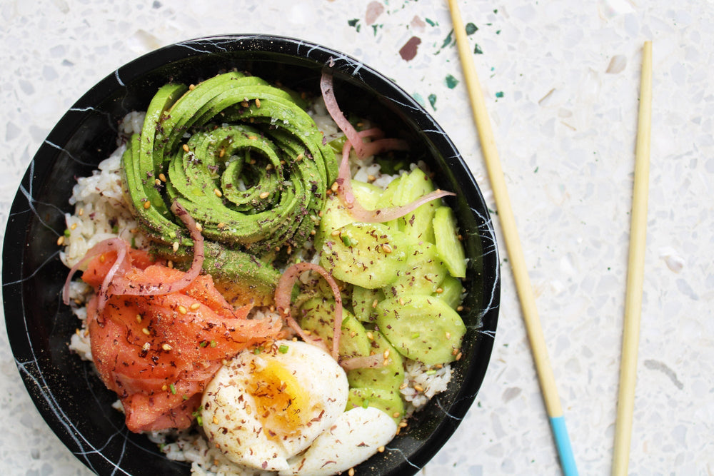 A bowl of rice with lox, hard boiled egg, cucumber slices and rolled avocado, all topped with Furikake seasoning.