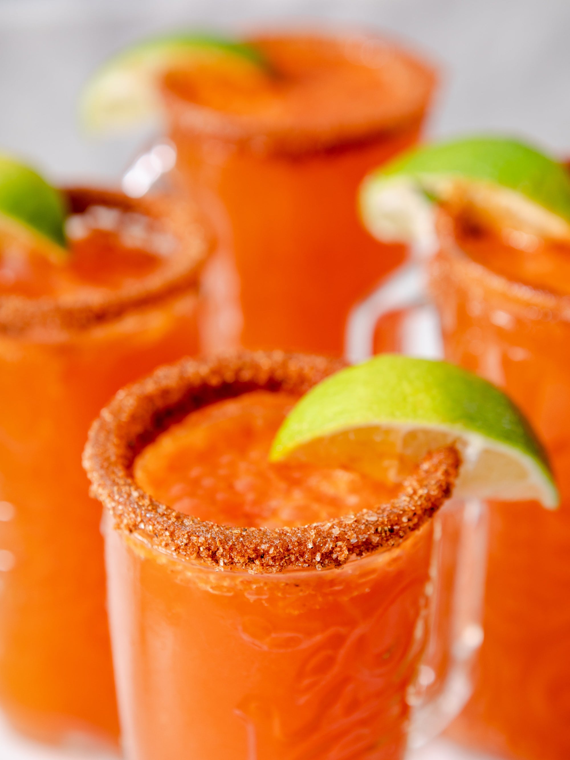 Four glasses filled with orange drinks, garnished with tajin and slices of lime.