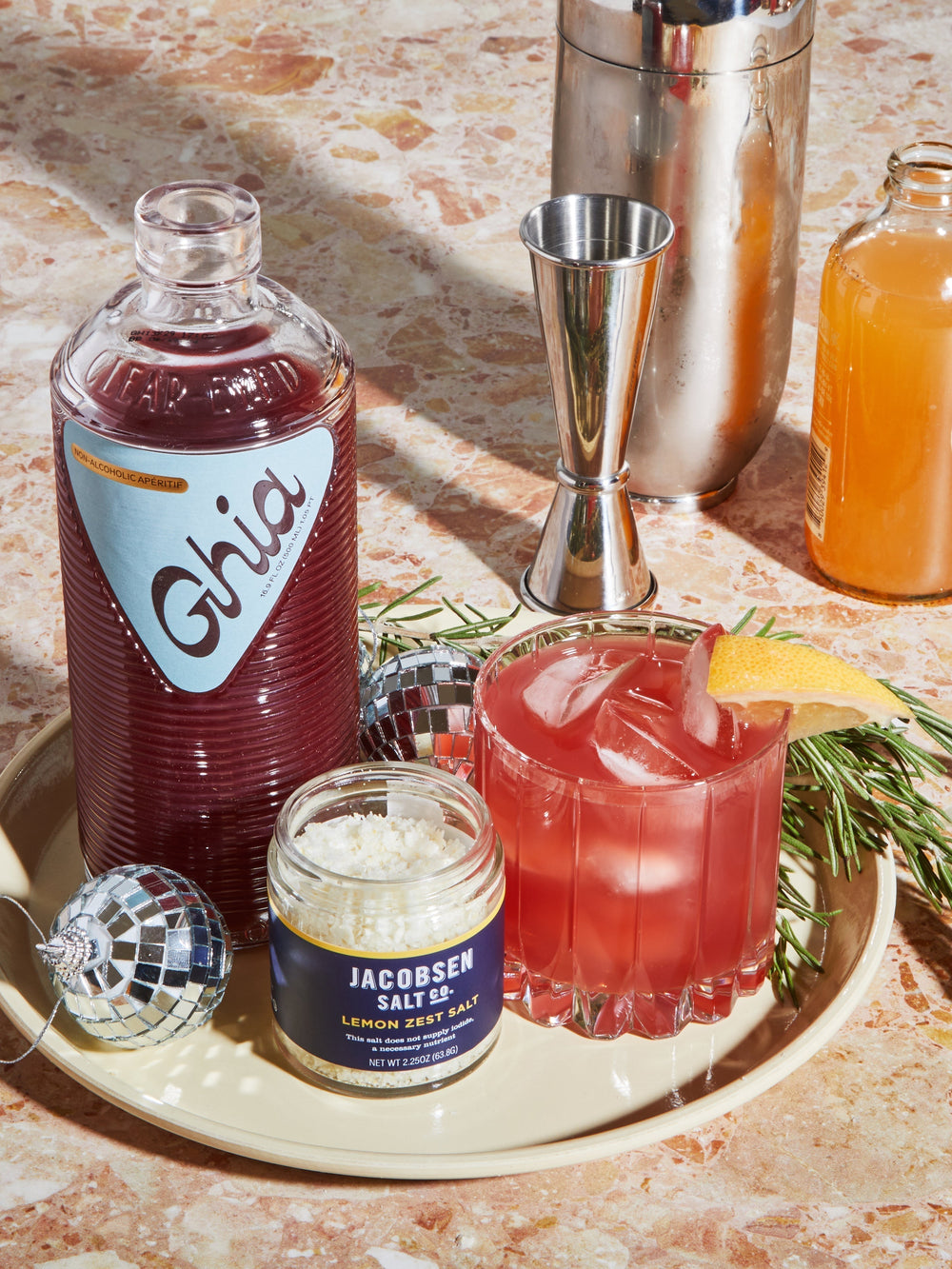 A bottle of Ghia aperitif, a jar of Jacobsen Infused Lemon Zest Salt, and a mocktail in a glass with a lemon wedge.