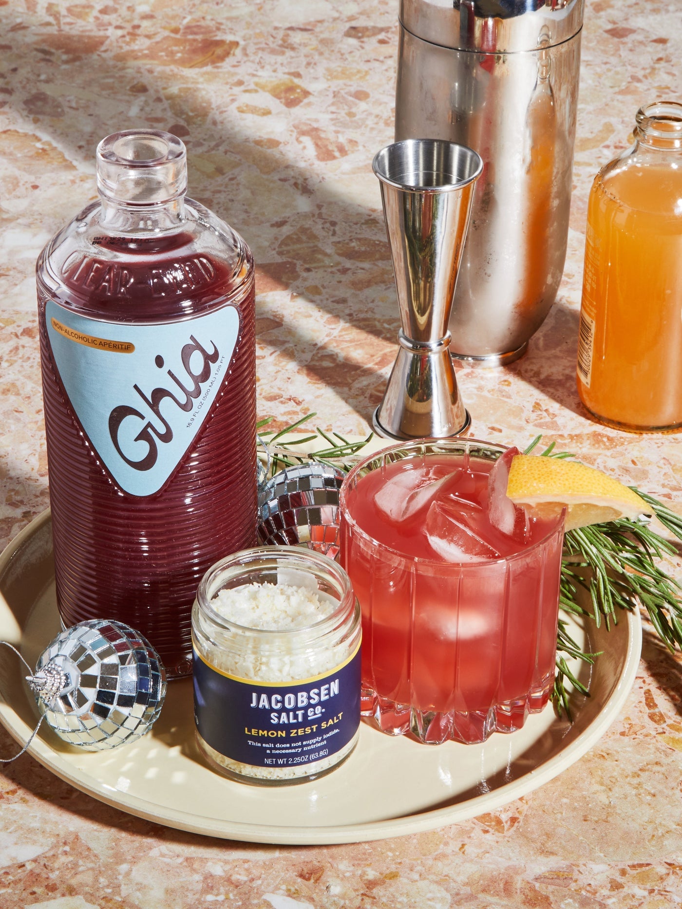 A bottle of Ghia aperitif, a jar of Jacobsen Infused Lemon Zest Salt, and a mocktail in a glass with a lemon wedge.