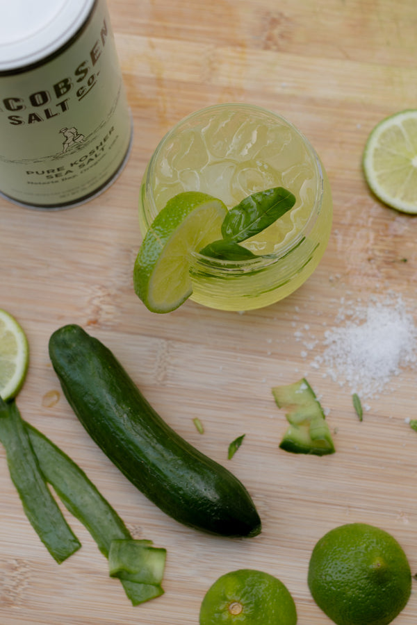 A green beverage with ice in a glass, garnished with lime. Sliced limes, a cucumber and Jacobsen Kosher Salt surround the glass.