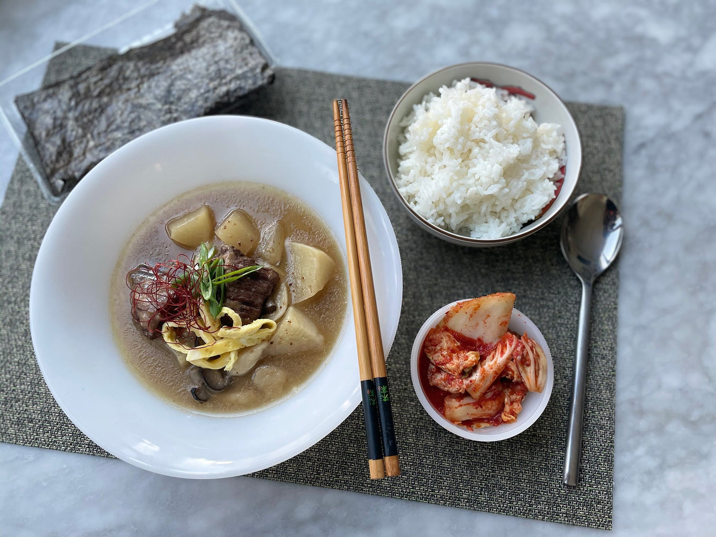 A bowl of soup with chopsticks on a placemat, with a bowl of rice and a bowl of kimchi to the right. 
