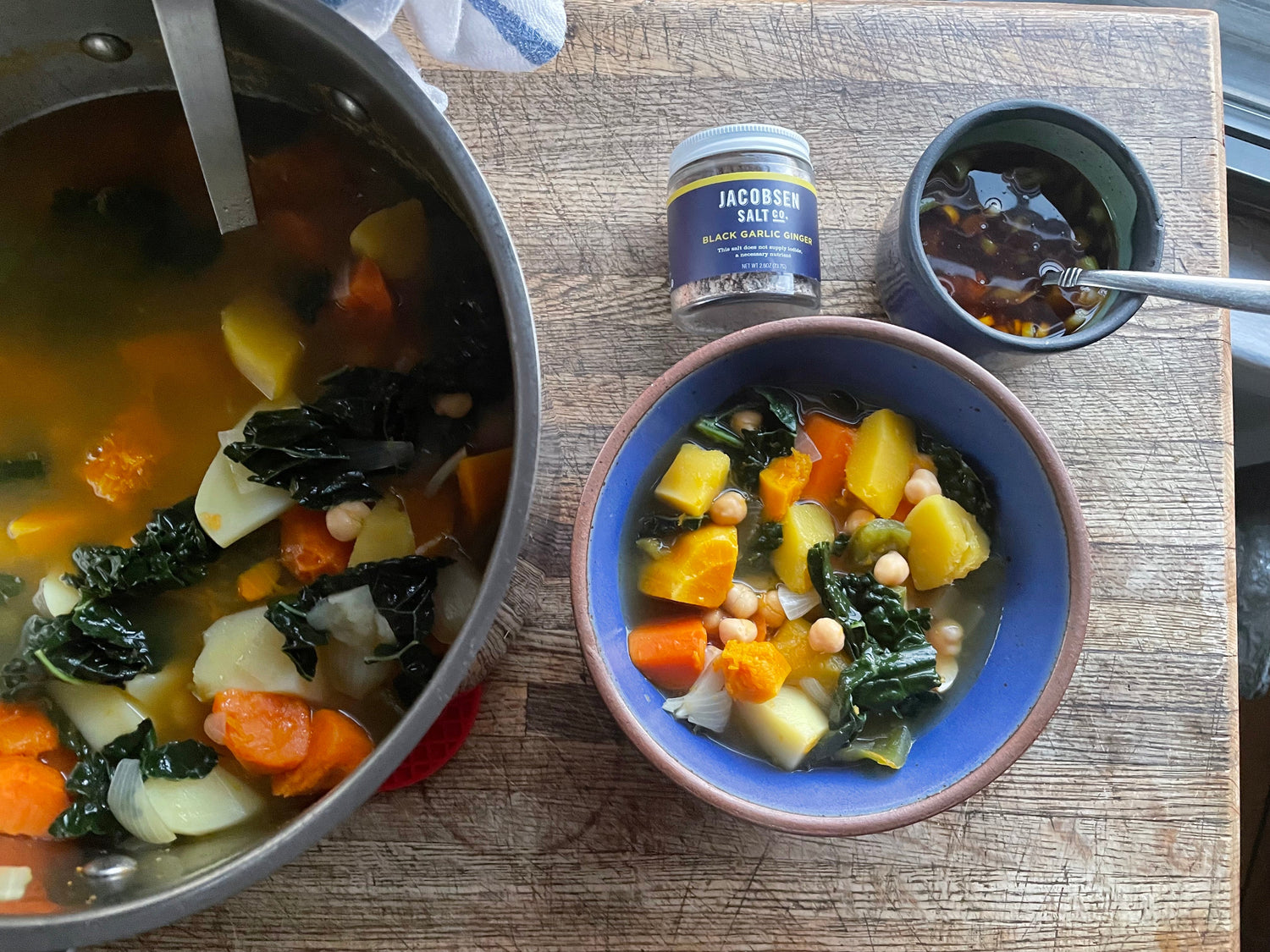 A pot and a bowl of vegetable stew on a wooden countertop. Next to the bowl is a jar of salt and a bowl of sauce.