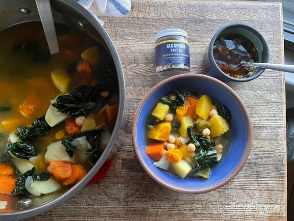 A pot and a bowl of vegetable stew on a wooden countertop. Next to the bowl is a jar of salt and a bowl of sauce.