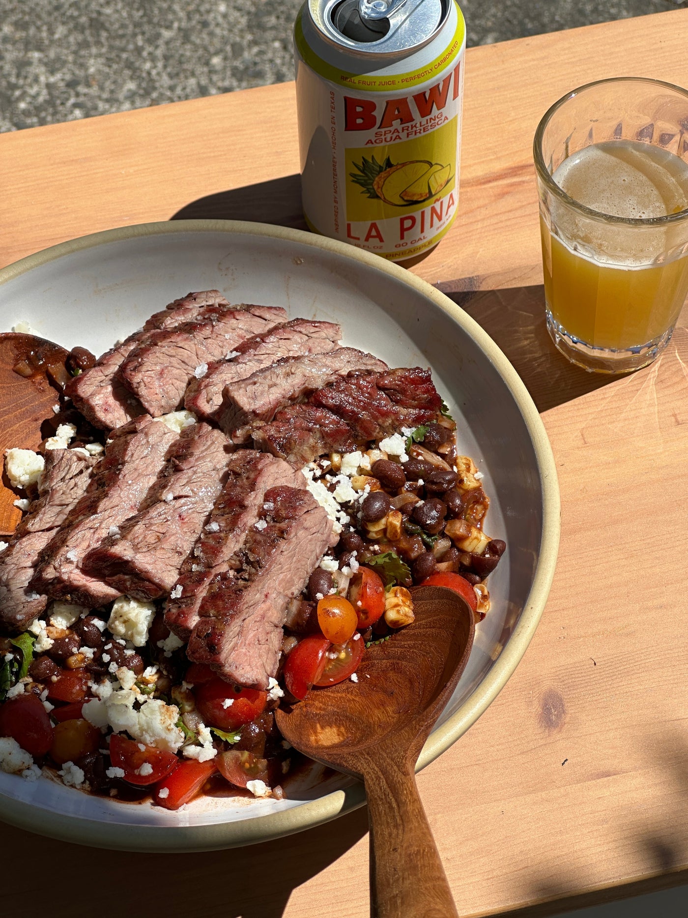 Slices of grilled steak on top of bean salad, next to an open can of Bawi and a glass containing the yellow beverage.