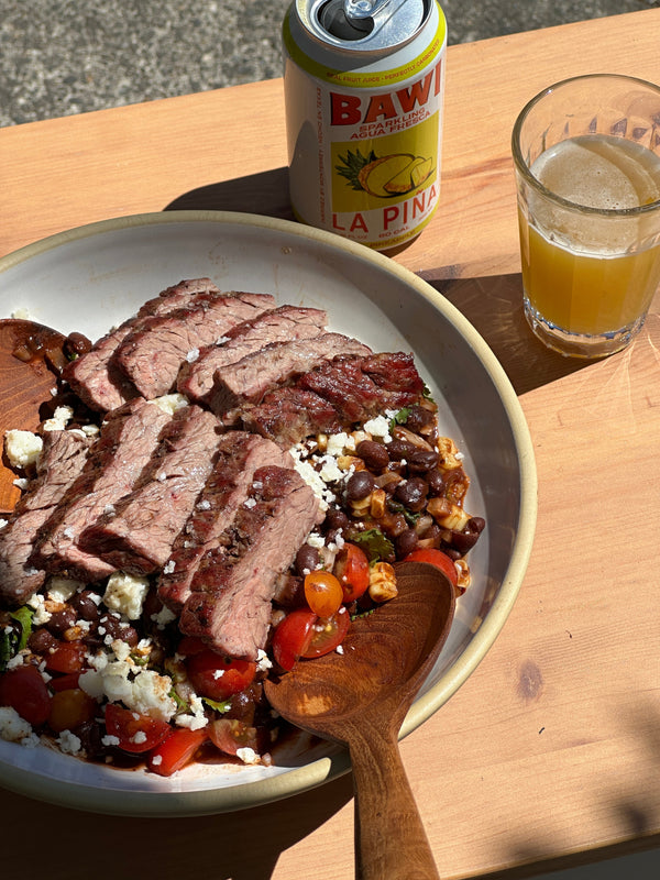 Slices of grilled steak on top of bean salad, next to an open can of Bawi and a glass containing the yellow beverage.