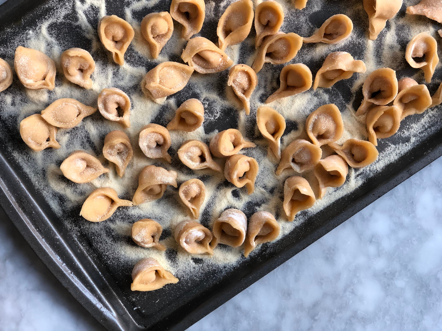 A baking sheet sits atop a granite surface. On top of the baking sheet are sweet potato tortellini which are covered in semolina flour.