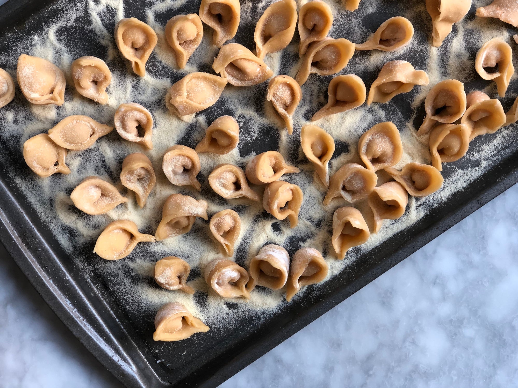 A baking sheet sits atop a granite surface. On top of the baking sheet are sweet potato tortellini which are covered in semolina flour.