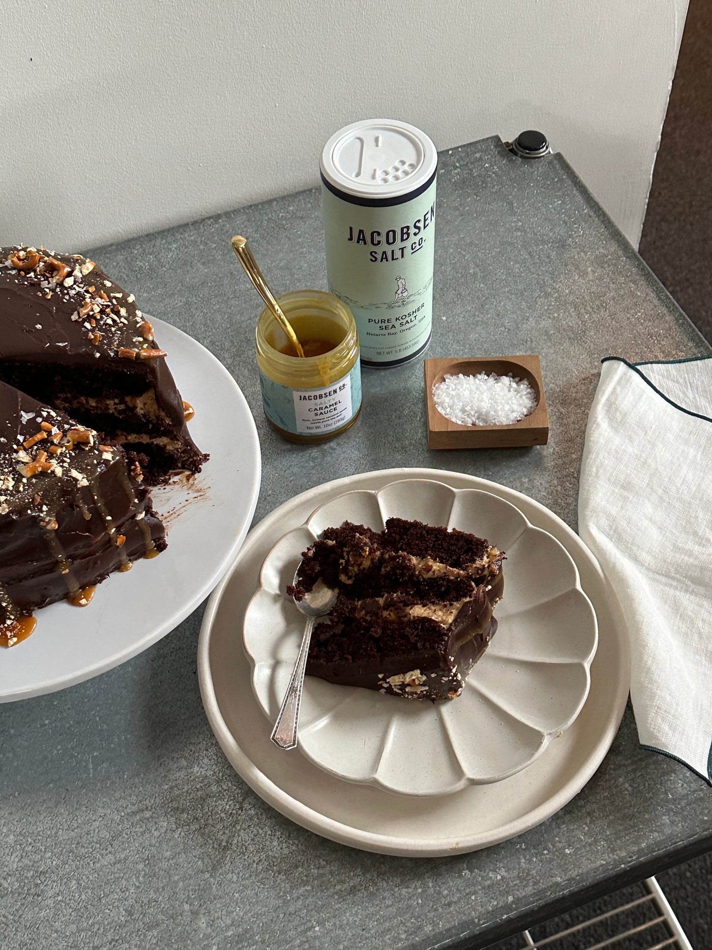Plated slice of decadent chocolate cake, next to canister of Jacobsen Kosher Sea Salt, Caramel Sauce, and a salt cellar.