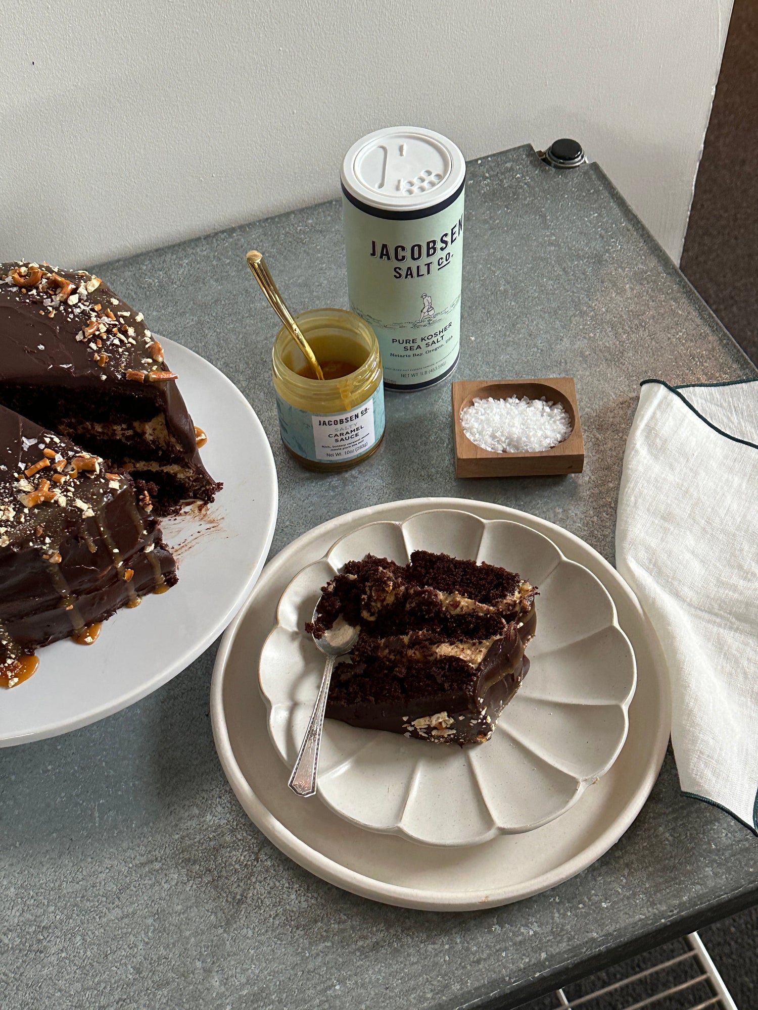 Plated slice of decadent chocolate cake, next to canister of Jacobsen Kosher Sea Salt, Caramel Sauce, and a salt cellar.