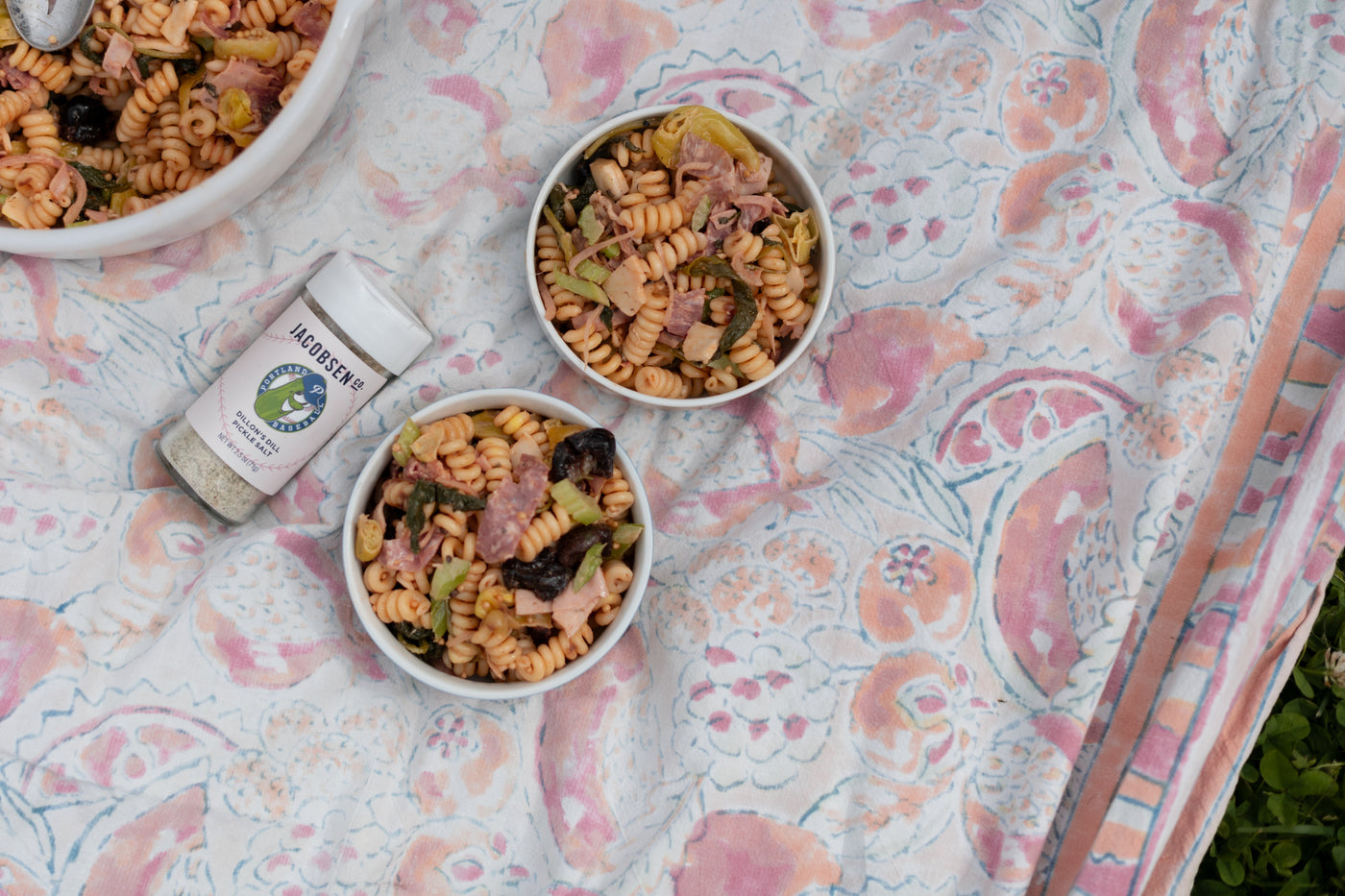 Bowls of pasta salad and a jar of pickle salt on a decorative tablecloth.
