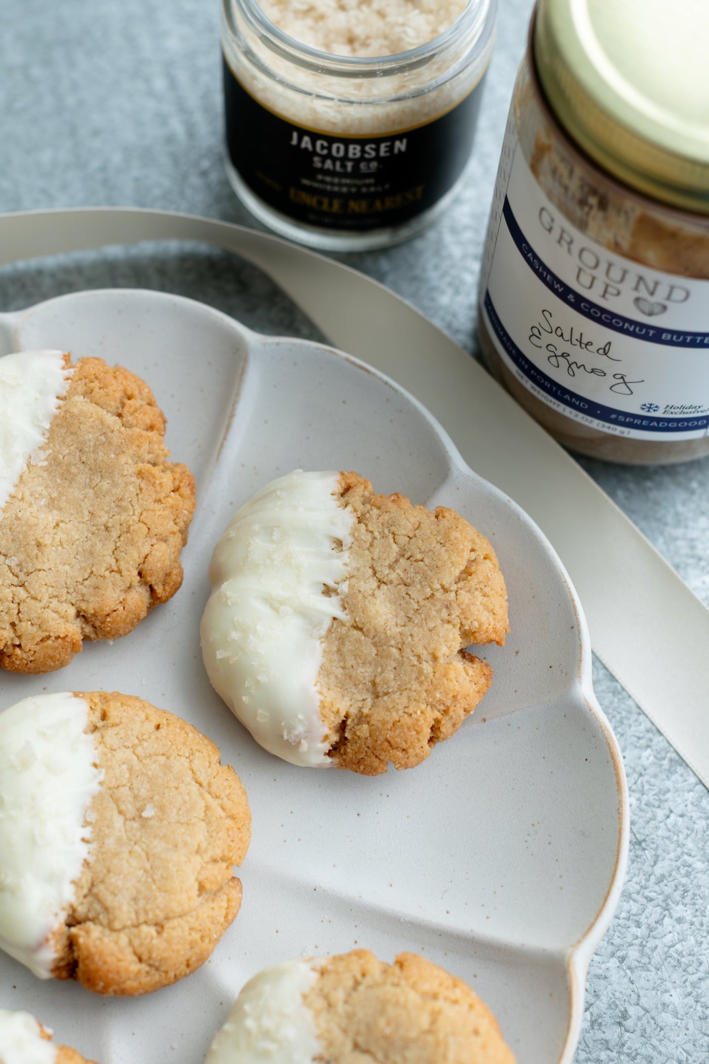 Round tan cookies, half dipped in white frosting on a plate, next to cherrywood smoked salt and salted eggnog spread.