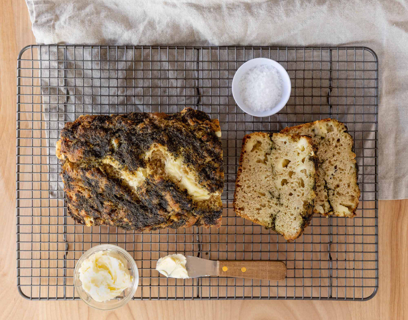 A cooling rack topped with a loaf of bread, a bowl of salt, and a bowl of soft butter with a spreading knife.