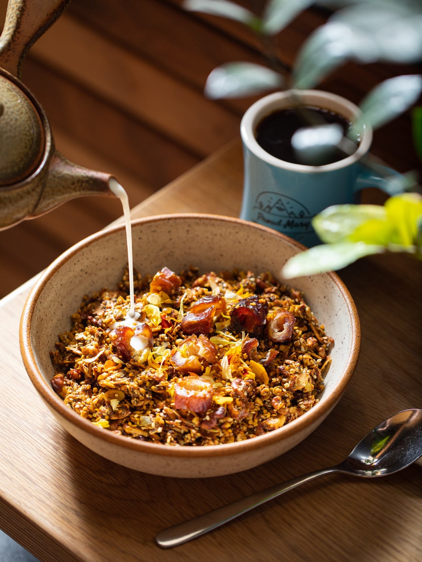 Milk is poured from a pitcher into a bowl of granola. A mug of coffee and a spoon are next to the bowl on a wooden table.