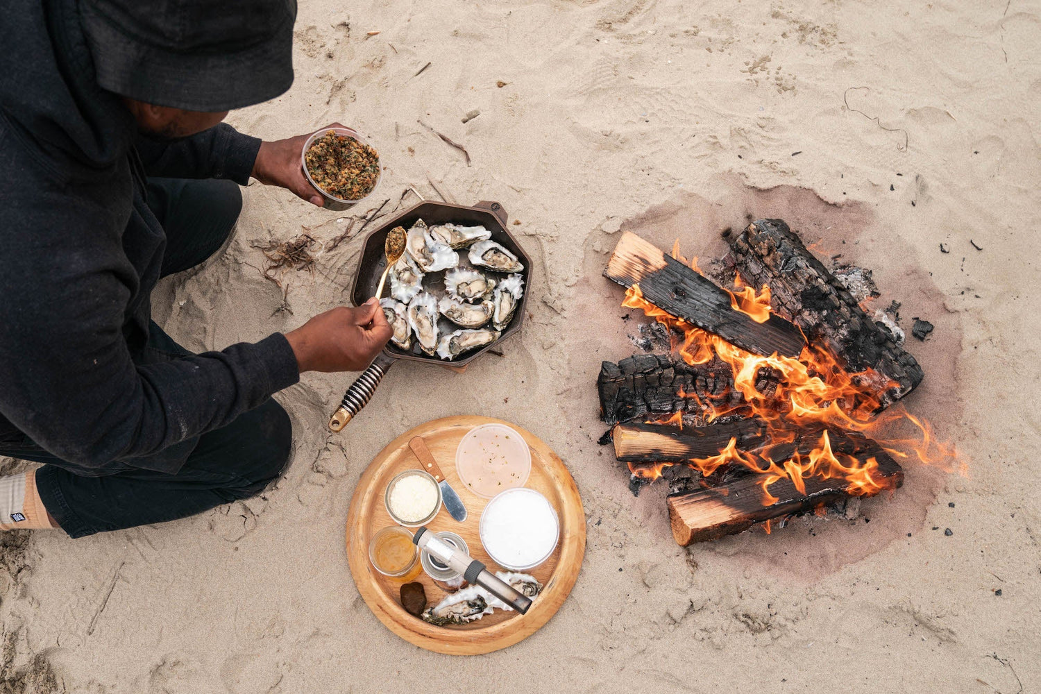 A person dresses a plate of raw oysters next to an open fire on the beach.