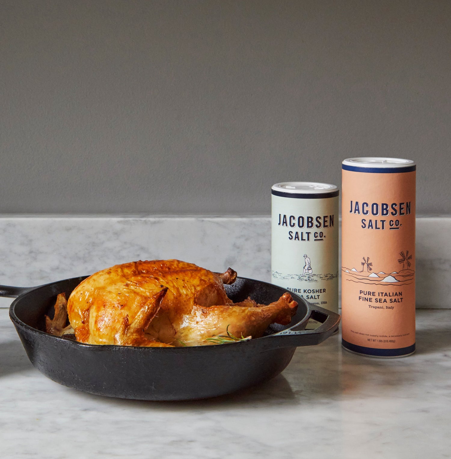 Two canisters of sea salt on a countertop and a roast chicken in a cast iron pan.