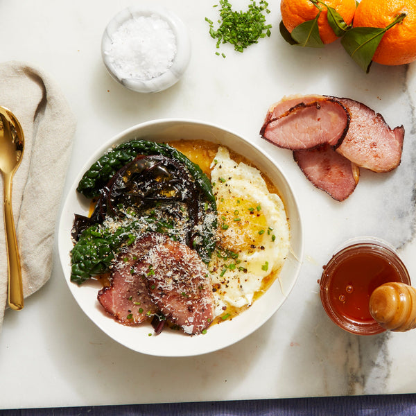 A bowl of savory porridge with greens on a marble countertop. Next to the bowl are slices of ham, a jar of honey, oranges, herbs, a bowl of salt, and a spoon.