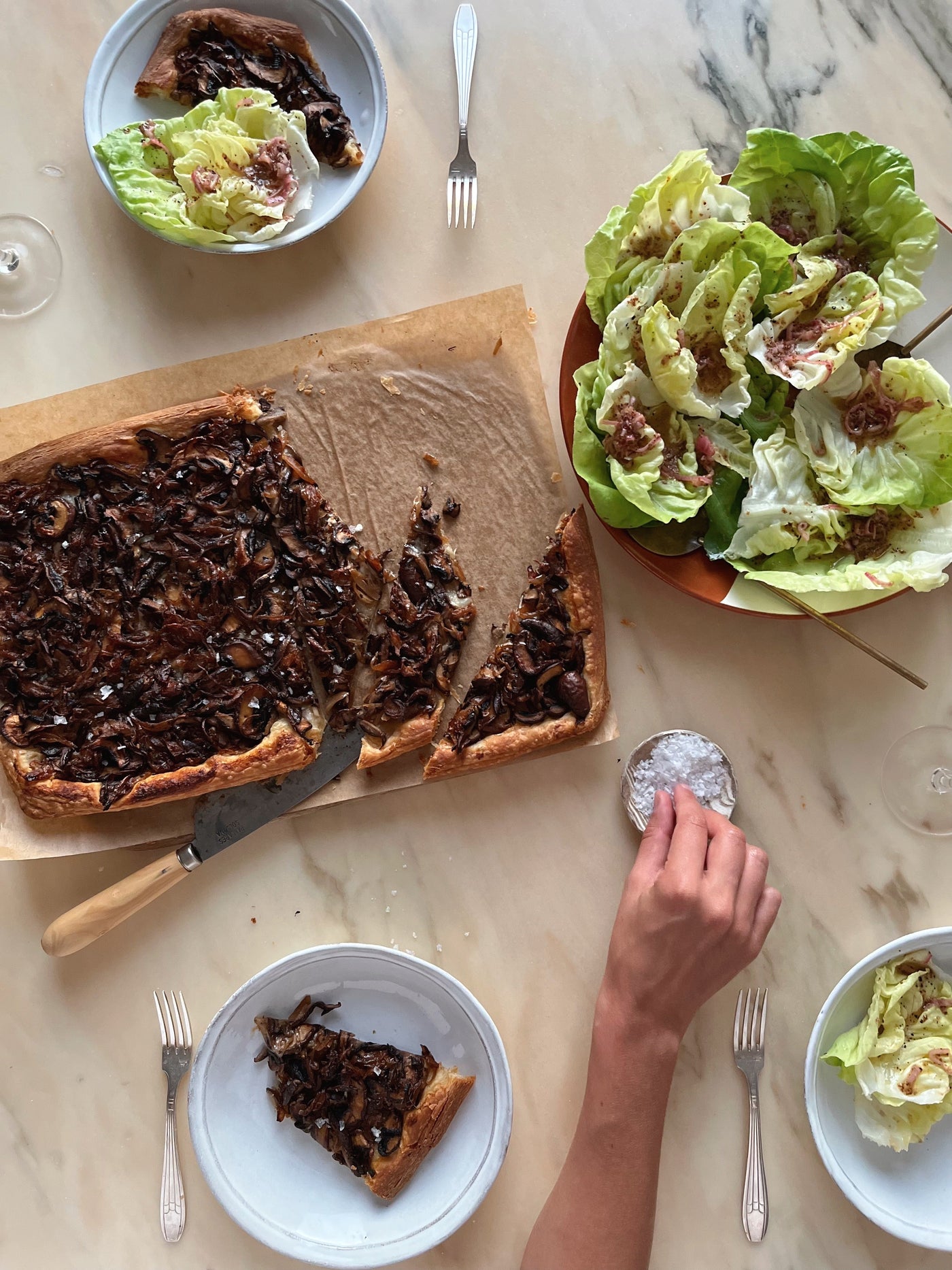 Plates of sliced mushroom galette and a fresh side salad. A person reaches for a pinch of salt on the table.