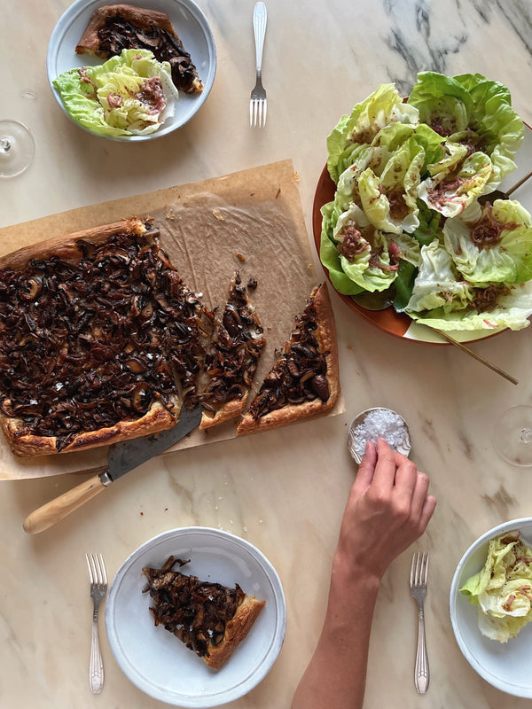 Plates of sliced mushroom galette and a fresh side salad. A person reaches for a pinch of salt on the table.
