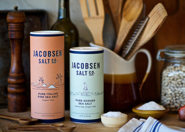 Jacobsen Pure Fine and Pure Kosher sea salt on countertop, with wooden utensils in the background.