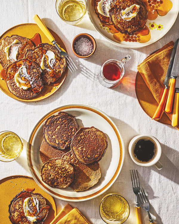 A breakfast table scape is laid out on a white tablecloth. There are pancakes on circular plates, jars of syrup, a bowl of salt, napkins, and cups on the tablecloth.