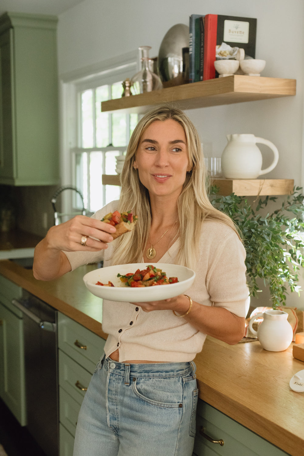 A woman stands in a kitchen and smiles. She holds a bowl in one hand, and food in the other.