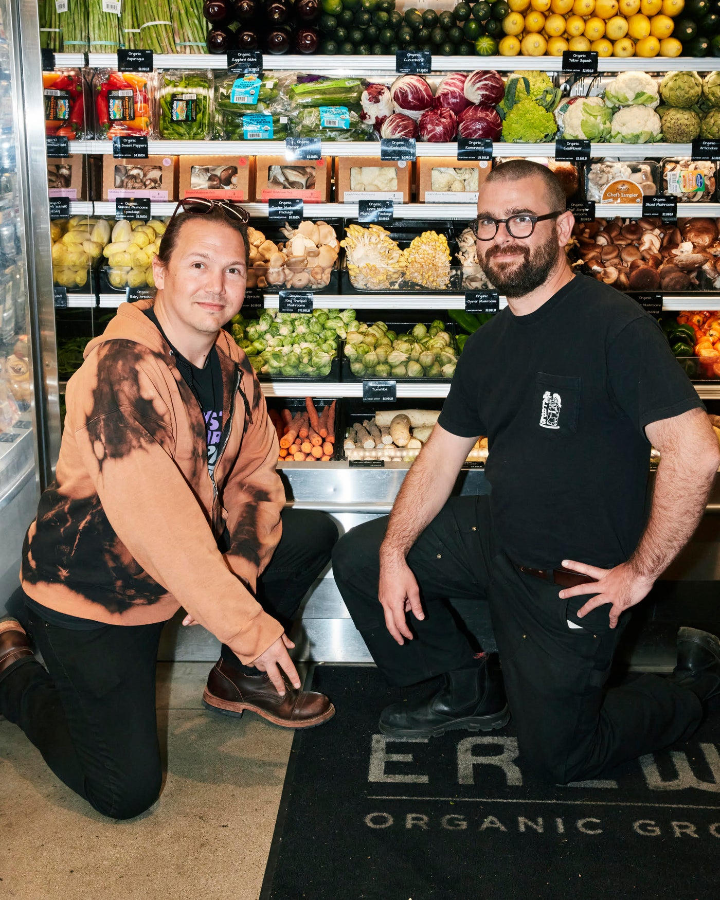 Two men kneel in the produce aisle of an Erewhon grocery store and smile at the camera.