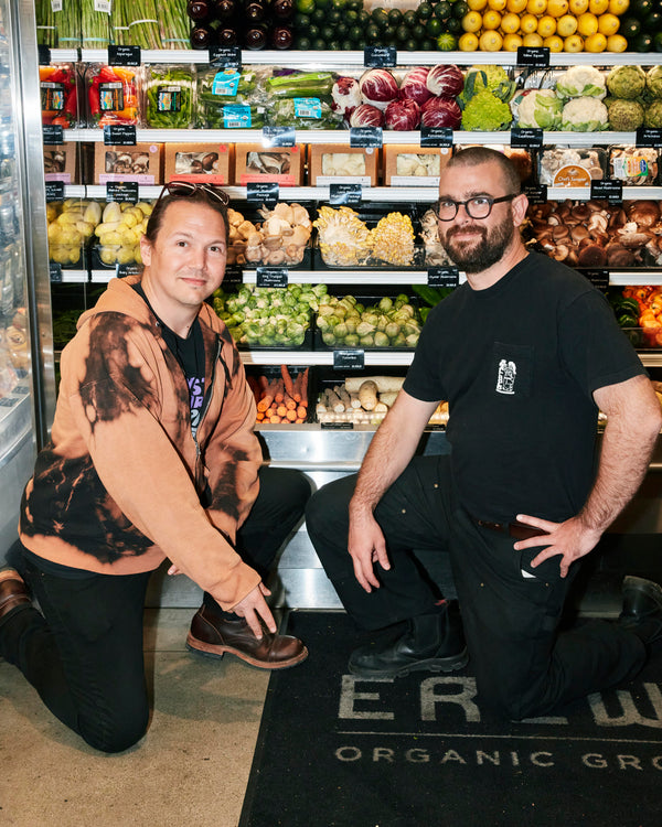 Two men kneel in the produce aisle of an Erewhon grocery store and smile at the camera.