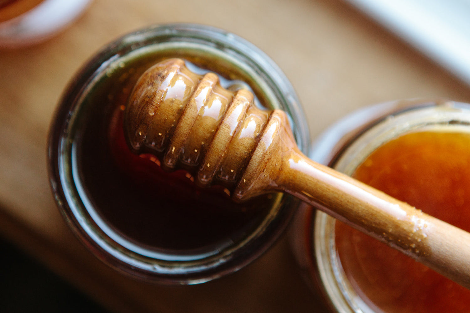 A wooden honey dipper with honey on it above an open jar of honey.
