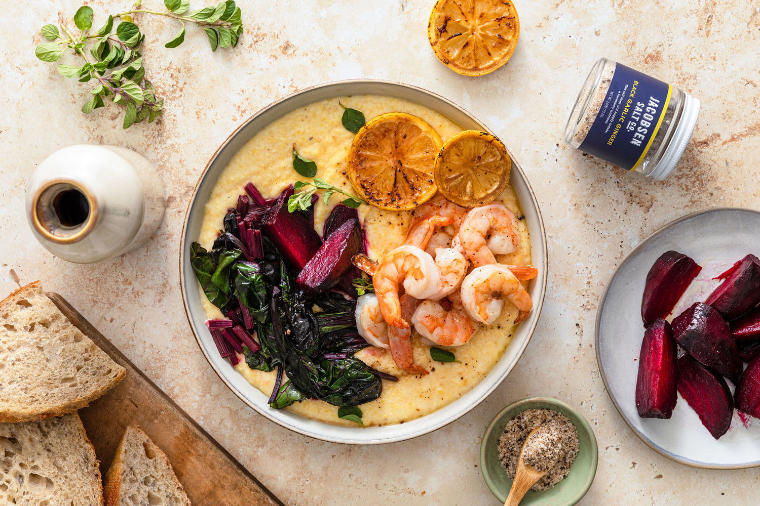 A bowl of polenta with greens, shrimp, and charred lemons on a granite countertop. Next to the bowl of polenta is a plate of beets, a jar of salt, charred lemon, and slices bread.