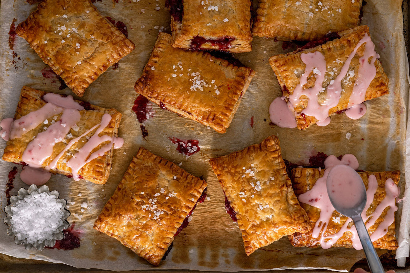 A spoon dribbles icing over freshly baked strawberry hand pies on a baking sheet.