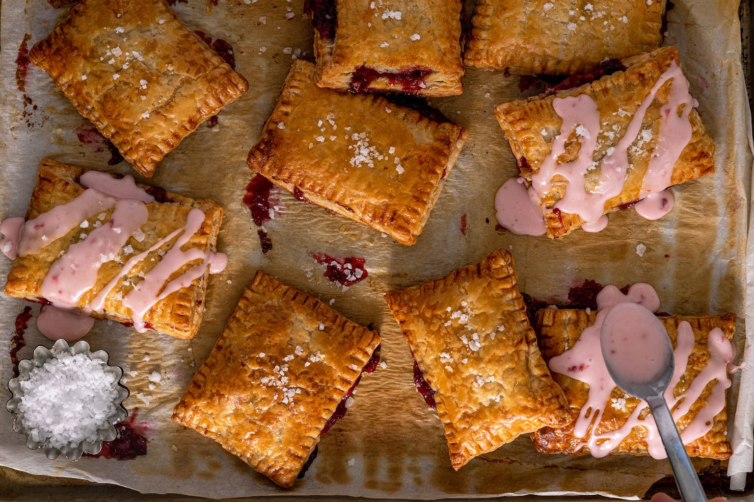 A spoon dribbles icing over freshly baked strawberry hand pies on a baking sheet.