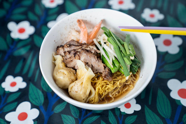 A bowl of noodles, meat and veggies on a flower-patterned table cloth.