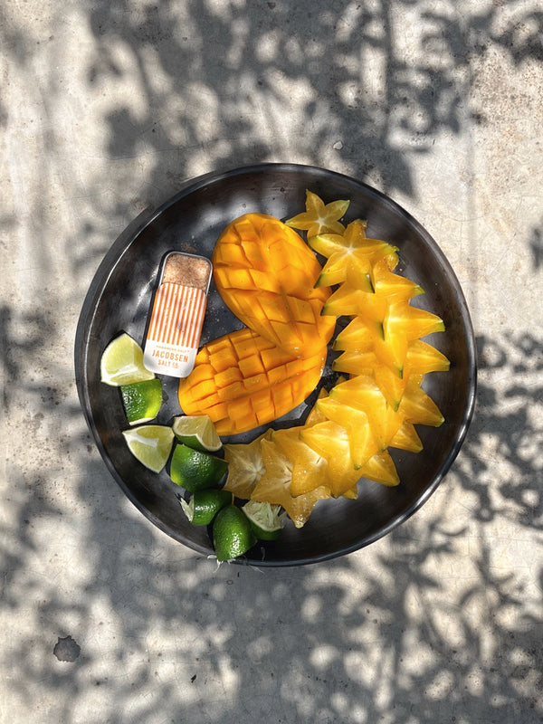 Sliced mangos, limes and starfruit on a plate with a Jacobsen Habanero Sea Salt Slide Tin. 
