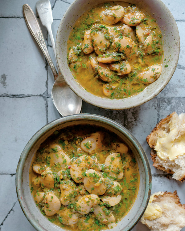 Two bowls of broth beans, crusty bread with butter, and two spoons on a tiled countertop.,