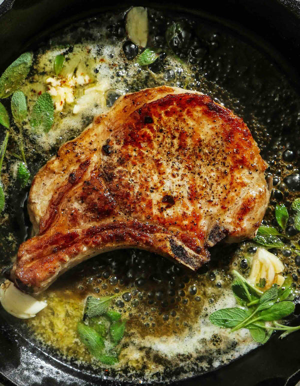 A pan-seared pork chop cooks in a cast-iron pan with herbs and butter.