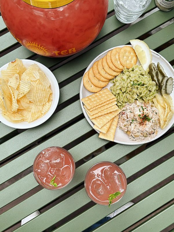 Plates of crackers, chips and toppings, next to glasses of pink iced beverage, all on top of a green slatted tabletop.
