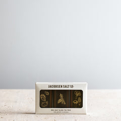 A rectangular box of Jacobsen Salt Co. Sea Salt Slide Tin Trio, featuring a cream-colored exterior and a window displaying three dark tins with gold botanical illustrations, set against a neutral light gray background.