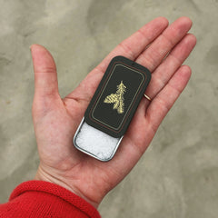 A hand holding a dark brown slide tin with a gold pine tree design on the lid, open and showing pure kosher salt.