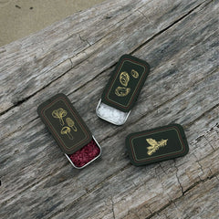 Three small rectangular dark brown metal tin decorated with gold botanical illustrations, arranged on a weathered wooden surface. One tin is open and filled with pink pinot noir salt, while the others show pure flake salt and pure kosher salt.