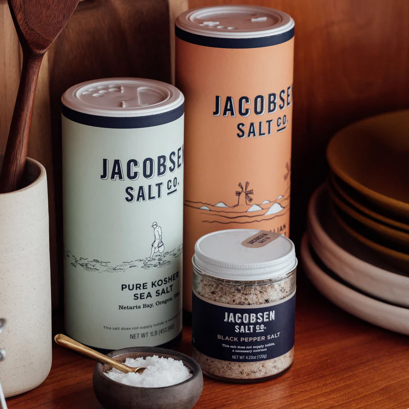 Two Jacobsen Salt Co. canisters on a shelf with a jar of black pepper salt and a bowl of flake salt.