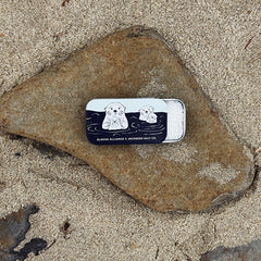 Small tin with sea otter illustration on a rock in the sand.