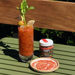 Jar of Jacobsen Sweet Hot Pepper Salt with a Bloody Mary on green picnic table. 